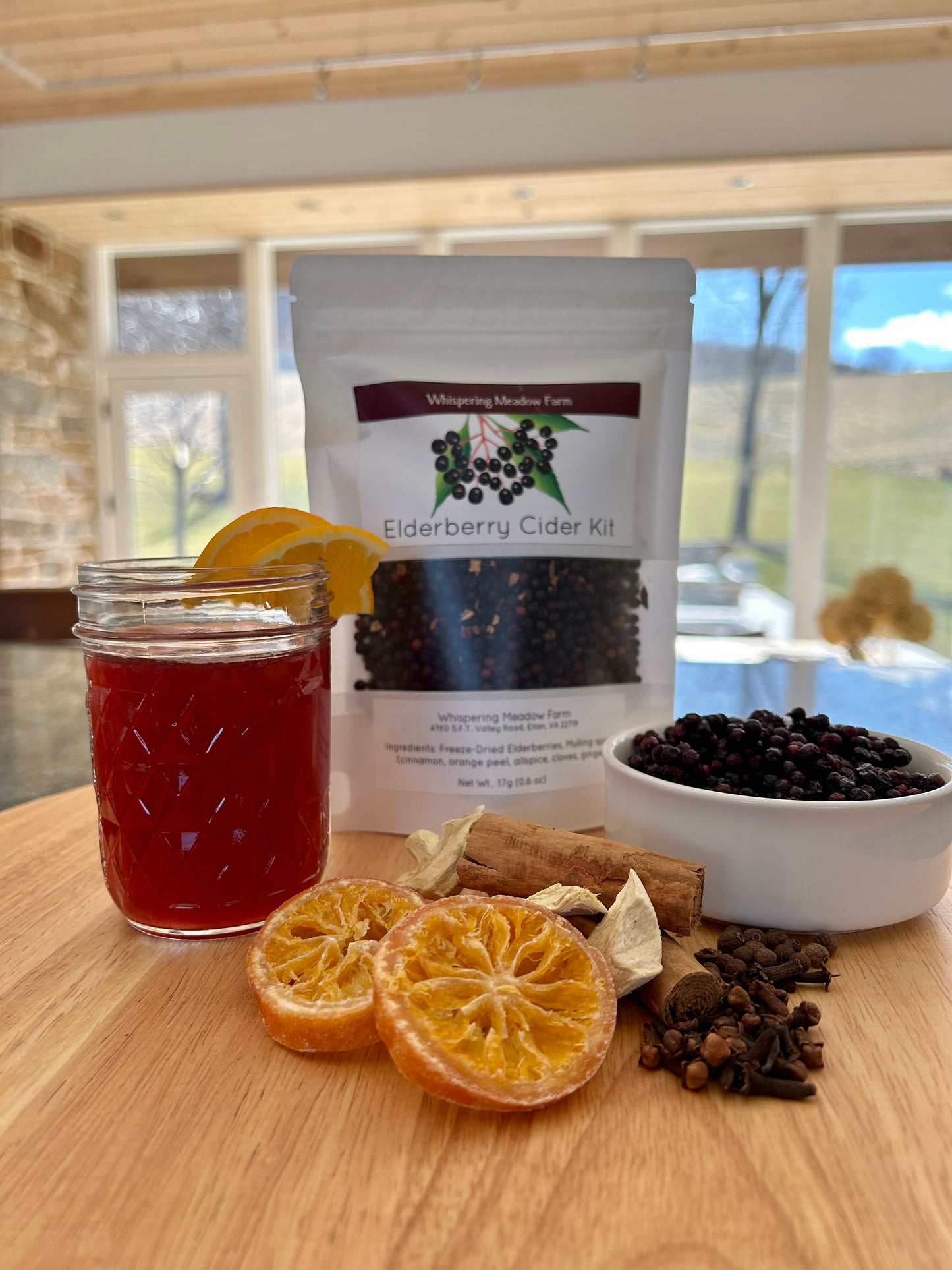 Bag of Elderberry Cider Kit placed among props like, a small bowl of elderberries, dried oranges, cinnamon cloves, allspice, and a small glass of the homemade cider.